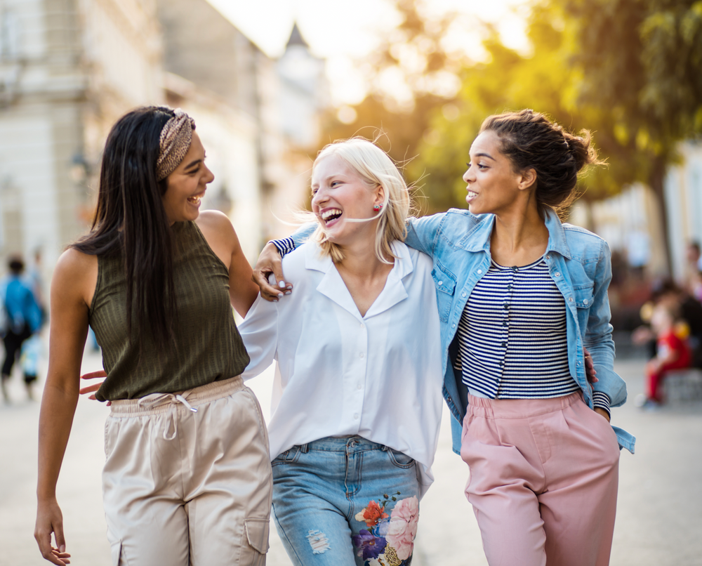 Three women together walking in a city with arms linked and smiling at each other